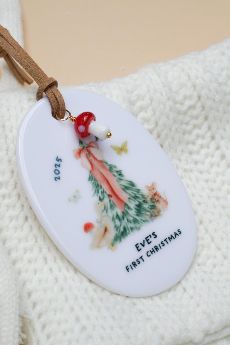 May include: White oval Christmas ornament with a painted Christmas tree design, butterflies, and the text "EVE'S FIRST CHRISTMAS 2025." A red and white mushroom bead hangs from a brown leather strap.