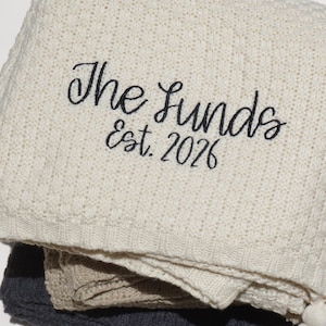 May include: Cream-colored knitted blanket with the embroidered text "The Lunds Est. 2026". The blanket is folded, revealing layers of gray and beige blankets. A white tassel is visible at the bottom.