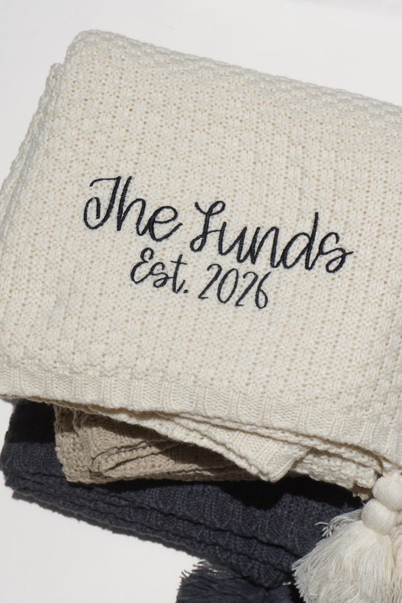 May include: Cream-colored knitted blanket with the embroidered text "The Lunds Est. 2026". The blanket is folded, revealing layers of gray and beige blankets. A white tassel is visible at the bottom.