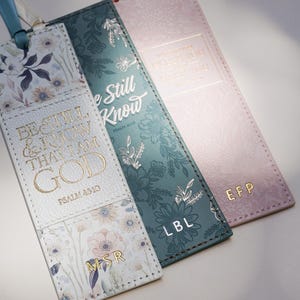 May include: Three leather bookmarks with floral designs and ribbon ties. One is white with gold lettering that reads "Be Still & Know That I Am God." Another is teal with "We Still Know" and "LBL." The third is pink with "EFP."