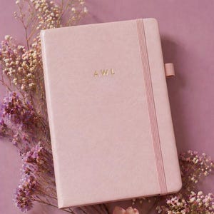 May include: A light pink notebook with a smooth cover featuring the gold embossed letters AWL in the center. The notebook includes a matching vertical elastic closure band and a small elastic pen loop on the right side, presented in a rectangular shape.