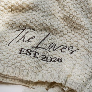 May include: Cream-colored knitted blanket with the embroidered text "The Loves EST. 2026". The blanket has a textured pattern and is finished with tassels along the edge, perfect for home decor.