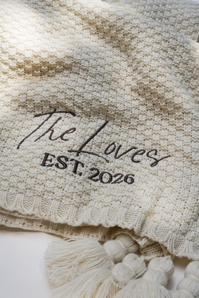 May include: Cream-colored knitted blanket with the embroidered text "The Loves EST. 2026". The blanket has a textured pattern and is finished with tassels along the edge, perfect for home decor.
