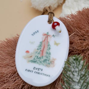 May include: White oval Christmas ornament with a Christmas tree design, pink bow, and the text "EVE'S FIRST CHRISTMAS 2025". The ornament has a brown leather cord and a mushroom charm.