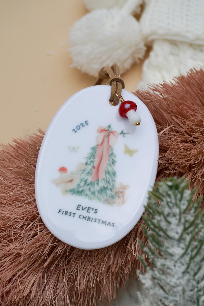 May include: White oval Christmas ornament with a Christmas tree design, pink bow, and the text "EVE'S FIRST CHRISTMAS 2025". The ornament has a brown leather cord and a mushroom charm.