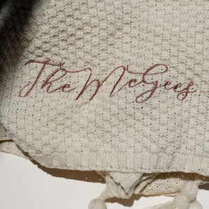 May include: Cream-colored knitted blanket with the embroidered text "The McGees" in a reddish-brown script. The blanket has a textured pattern and is finished with two cream-colored tassels.