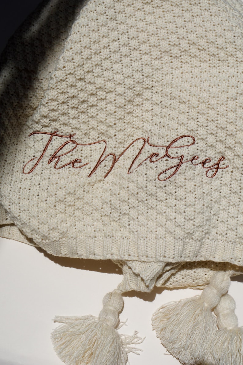 May include: Cream-colored knitted blanket with the embroidered text "The McGees" in a reddish-brown script. The blanket has a textured pattern and is finished with two cream-colored tassels.