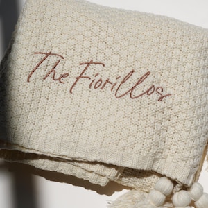 May include: Cream-colored knitted blanket with the embroidered text "The Fiorillos" in a brown script. The blanket has a textured pattern and is adorned with cream-colored tassels.