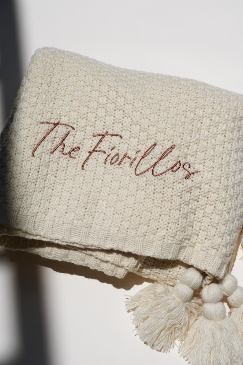 May include: Cream-colored knitted blanket with the embroidered text "The Fiorillos" in a brown script. The blanket has a textured pattern and is adorned with cream-colored tassels.