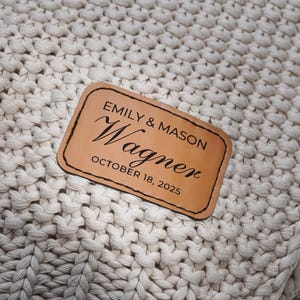 May include: A close-up of a cream-colored, chunky knit blanket. A rectangular leather patch is sewn onto the blanket, with the names "Emily & Mason Wagner" and the date "October 18, 2025" printed on it.