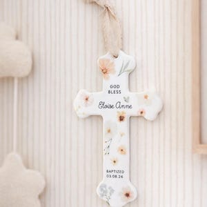 May include: A white ceramic cross ornament featuring a delicate floral pattern in soft pastel tones. The text reads GOD BLESS, Eloise Anne, and BAPTIZED 03.08.26. A frayed beige fabric ribbon is attached to the top for hanging purposes.