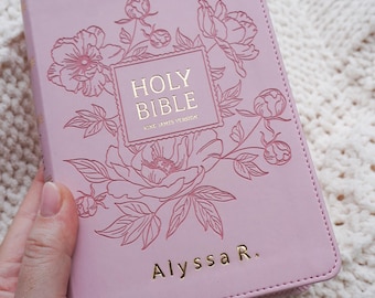 Puede incluir: Una Santa Biblia rosa con ilustraciones florales y letras doradas. La portada presenta las palabras "Holy Bible" y "King James Version" dentro de un cuadrado, rodeadas de diseños de flores y hojas. El nombre "Alyssa R." también está en oro.