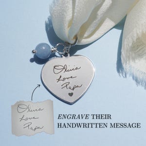 May include: A silver heart-shaped pendant engraved with the words "Olivia Love Papa" and a small heart. The pendant is attached to a white scarf and a light blue bead. The text "ENGRAVE THEIR HANDWRITTEN MESSAGE" is also visible.