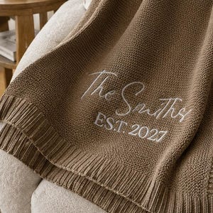May include: A brown knitted throw blanket featuring a ribbed border and white embroidered text that reads The Smiths EST. 2027. The fabric has a consistent textured knit pattern throughout, showcasing the personalized lettering in a script font.