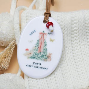 May include: Oval Christmas ornament with a painted Christmas tree design, the text "EVE'S FIRST CHRISTMAS" and the year 2025. The ornament has a brown leather strap and a red and white mushroom charm.