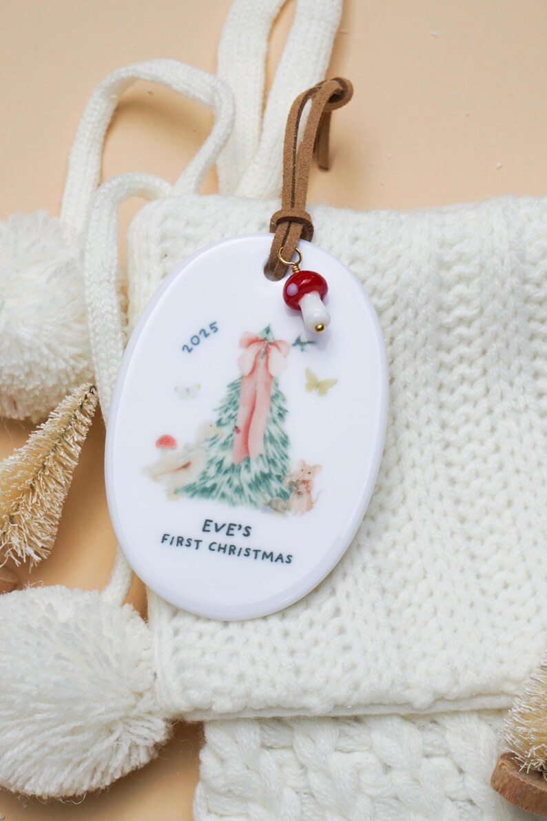 May include: Oval Christmas ornament with a painted Christmas tree design, the text "EVE'S FIRST CHRISTMAS" and the year 2025. The ornament has a brown leather strap and a red and white mushroom charm.