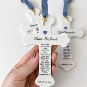 May include: A white ceramic cross ornament featuring a blue heart, a gold cross charm, and a blue fabric ribbon. The text reads Oliver Newland, The Lord bless you and protect you, The Lord be kind and gracious to you, and February 12, 2026.