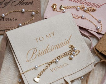 May include: Gold-colored pearl bracelets with heart-shaped charms are displayed on small, off-white and pink pouches. The pouches are printed with the phrases "To My Bridesmaid" and "To My Maid of Honor" in gold lettering. The bracelets are from VUN&Co.