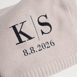 May include: A light beige knit blanket featuring black embroidered text. The embroidery displays the letters K and S separated by a vertical line, with the date 8.8.2026 positioned directly underneath the letters in a clean, dark font.