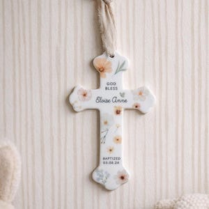May include: A white ceramic cross ornament featuring a delicate floral pattern in soft pastel tones. The text reads "GOD BLESS", "Eloise Anne", and "BAPTIZED 03.08.26". A frayed beige fabric ribbon is attached to the top for hanging purposes.