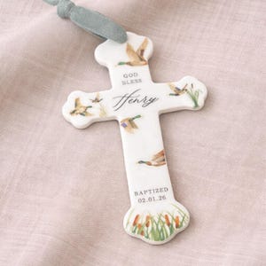 May include: A white ceramic cross ornament featuring watercolor illustrations of ducks and cattails. The text reads GOD BLESS, Henry, and BAPTIZED 02.01.26. A sage green velvet ribbon is attached to the top for hanging, creating a decorative keepsake.
