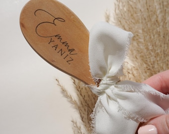 May include: Wooden oval-shaped name tag with the name "Emma Yaniz" in cursive script. A white fabric bow is tied to the tag. The background features dried ornamental grass.