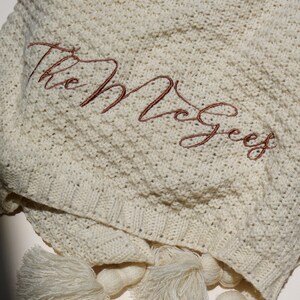 May include: Cream-colored knitted blanket with the embroidered name "The McGees" in a reddish-brown script. The blanket has a textured pattern and is finished with cream-colored tassels.