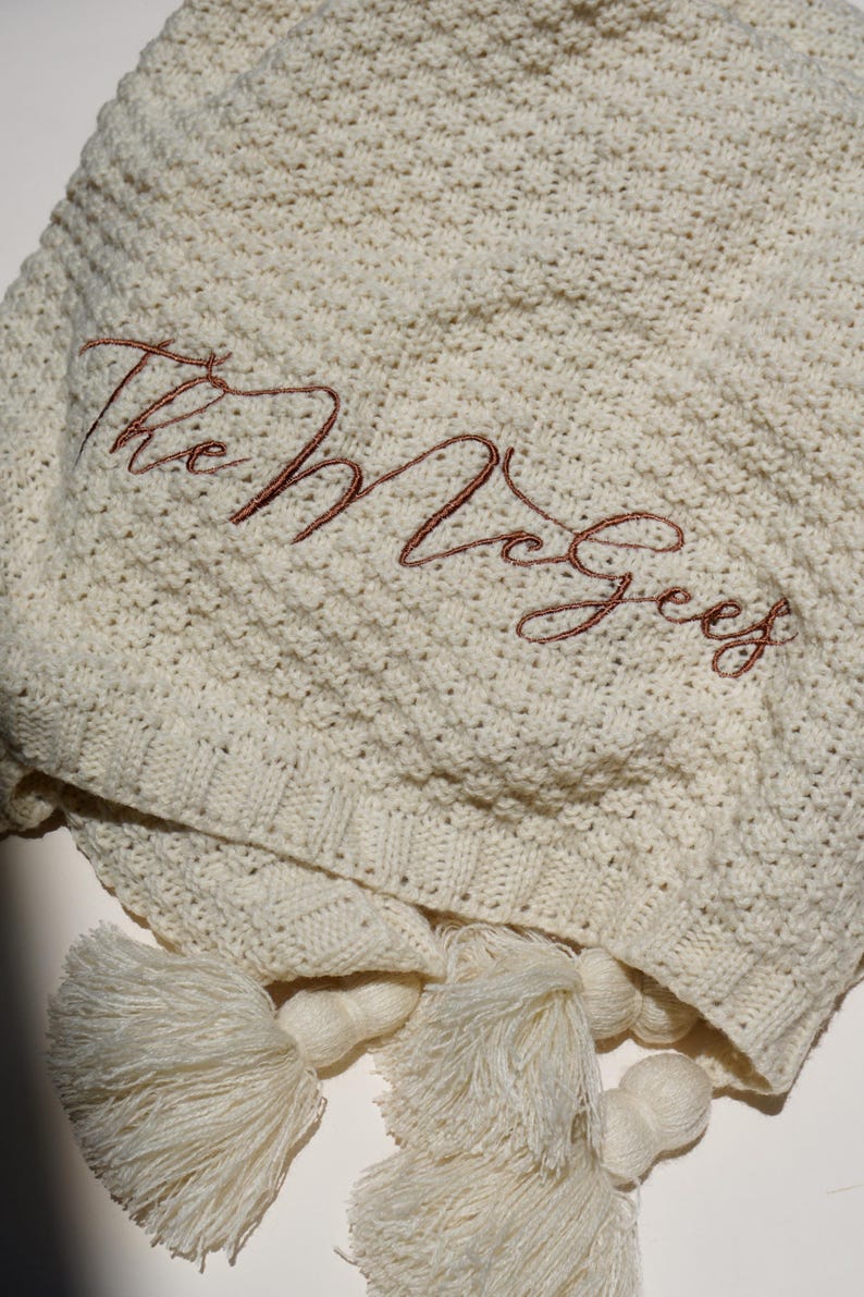 May include: Cream-colored knitted blanket with the embroidered name "The McGees" in a reddish-brown script. The blanket has a textured pattern and is finished with cream-colored tassels.