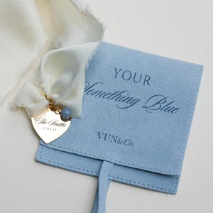 May include: A light blue felt pouch with the text "YOUR Something Blue" and a gold heart-shaped charm. The charm is engraved with "The Smiths" and the date 11.05.25. A white ribbon is attached to the charm and pouch.