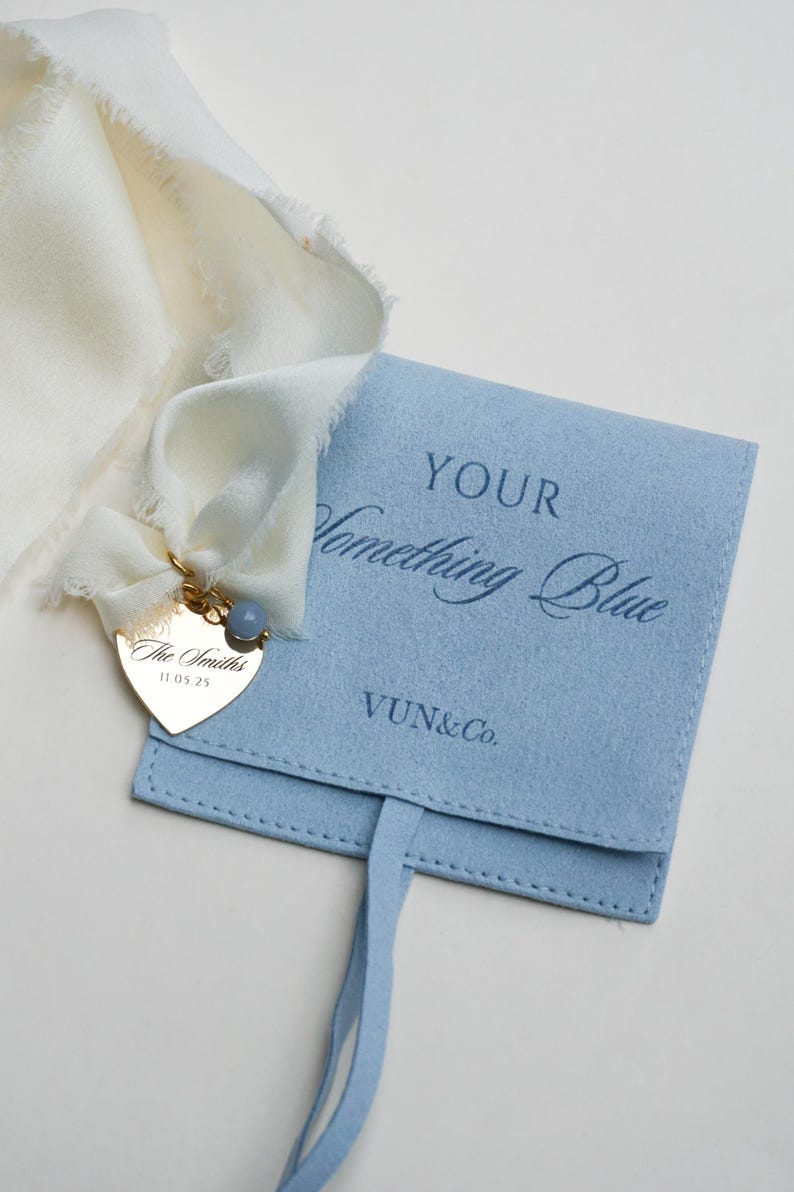 May include: A light blue felt pouch with the text "YOUR Something Blue" and a gold heart-shaped charm. The charm is engraved with "The Smiths" and the date 11.05.25. A white ribbon is attached to the charm and pouch.