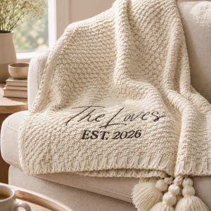 May include: Cream-colored knitted throw blanket with the text "The Loves EST. 2026" embroidered in black. The blanket has a textured pattern and is draped over a beige sofa. Tassels hang from the edge.