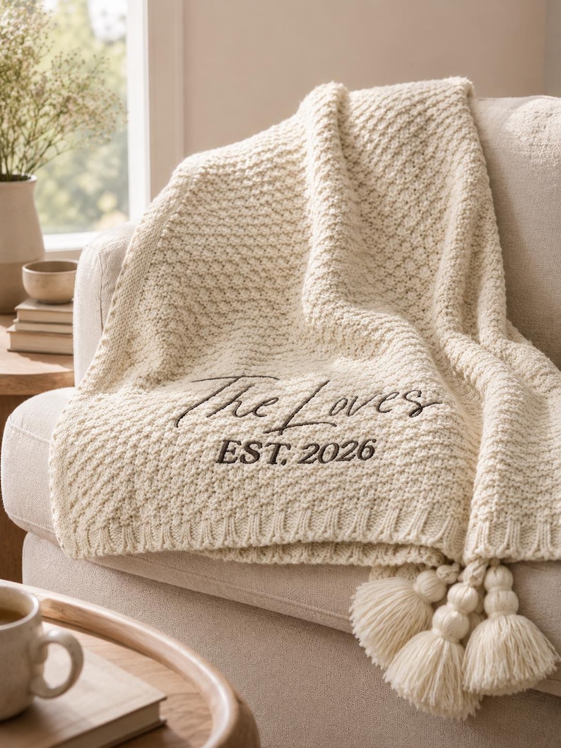 May include: Cream-colored knitted throw blanket with the text "The Loves EST. 2026" embroidered in black. The blanket has a textured pattern and is draped over a beige sofa. Tassels hang from the edge.