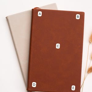 May include: Two leather-bound journals, one tan and one brown, are displayed on a white surface. The brown journal is open, revealing a tan interior. The text "CHOOSE PLACEMENT" is at the top, and the brand name is at the bottom.