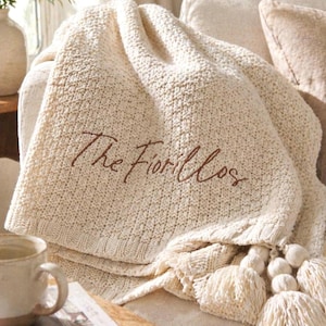 May include: Cream-colored knitted throw blanket with the embroidered text "The Fiorillos". The blanket has a textured pattern and is adorned with tassels and pom-poms. The blanket is draped over a sofa, creating a cozy and inviting atmosphere.