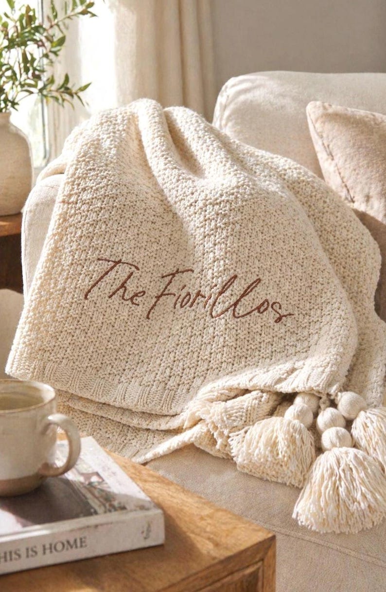 May include: Cream-colored knitted throw blanket with the embroidered text "The Fiorillos". The blanket has a textured pattern and is adorned with tassels and pom-poms. The blanket is draped over a sofa, creating a cozy and inviting atmosphere.