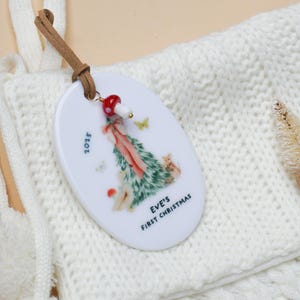 May include: Oval Christmas ornament with a forest scene, butterflies, and the text "EVE'S FIRST CHRISTMAS" and "2025". The ornament has a brown leather strap and a red and white mushroom charm.