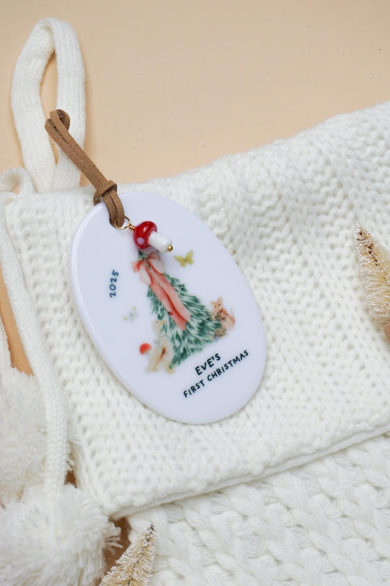 May include: Oval Christmas ornament with a forest scene, butterflies, and the text "EVE'S FIRST CHRISTMAS" and "2025". The ornament has a brown leather strap and a red and white mushroom charm.