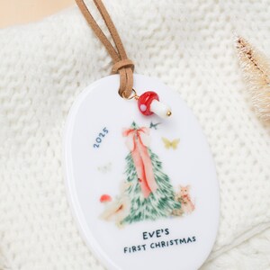 May include: White ceramic Christmas ornament with a brown leather cord. The ornament features a Christmas tree design with a pink bow, the text "EVE'S FIRST CHRISTMAS" and the year 2025. A red mushroom charm is attached.