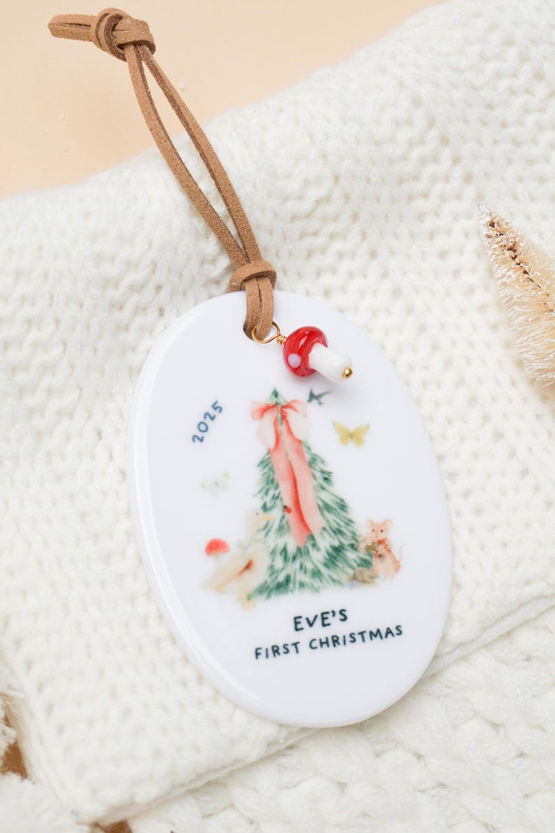 May include: White ceramic Christmas ornament with a brown leather cord. The ornament features a Christmas tree design with a pink bow, the text "EVE'S FIRST CHRISTMAS" and the year 2025. A red mushroom charm is attached.