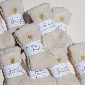 May include: A set of fuzzy white socks with gold heart embellishments and personalized labels for a wedding party. The labels include the names "Kayleigh Bridesmaid", "Marina Maid of Honor", "Victoria Bridesmaid", "Stacy Bride", "Jennifer Bridesmaid", "Lindsey Bridesmaid", "Candice Bridesmaid", and "Bri"