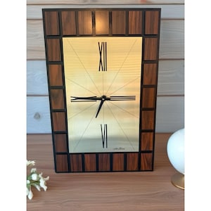 May include: Rectangular wall clock with a gold face and black hands. The clock face features Roman numerals and radial lines. The frame is made of dark wood blocks with a black border. The clock is on a wooden surface.