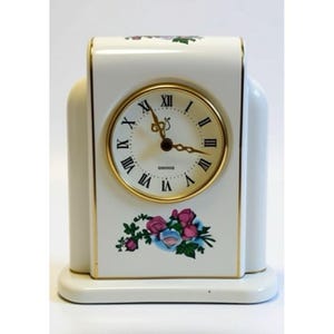 May include: A white ceramic mantel clock with a gold-tone bezel and Roman numeral clock face. The clock is decorated with floral accents in pink, blue, and green. The clock measures approximately 6 inches tall.