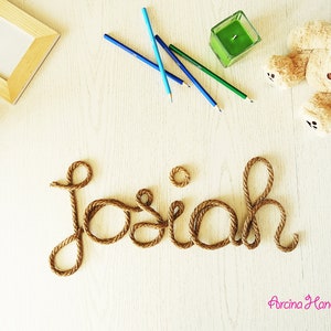 May include: A decorative name sign spelling the word Josiah in cursive script made from thick brown twisted rope. The display includes a light wood frame with the word love, three colored pencils, a green candle, and a plush teddy bear.