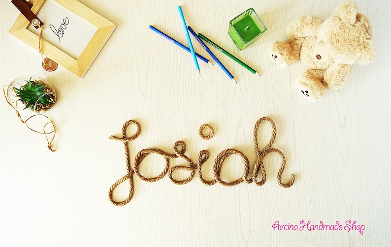 May include: A decorative name sign spelling the word Josiah in cursive script made from thick brown twisted rope. The display includes a light wood frame with the word love, three colored pencils, a green candle, and a plush teddy bear.