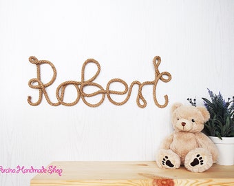 May include: A wall-mounted name sign spelling the name Robert, crafted from thick, twisted brown rope. The letters are connected in a continuous cursive script. A plush beige teddy bear and a small potted plant are positioned below the sign.