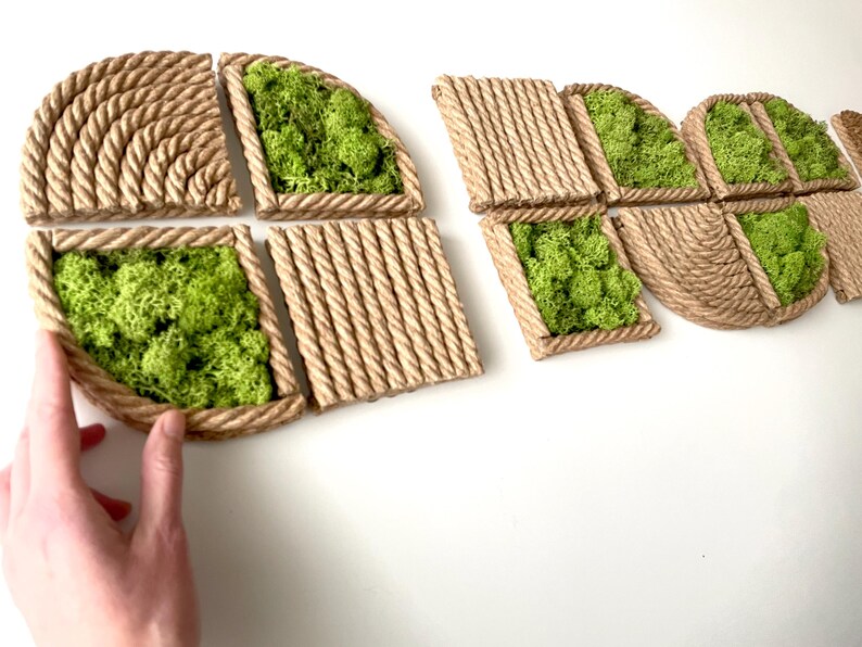 May include: A series of geometric wall art panels featuring natural green preserved moss encased in frames made of thick, twisted tan rope. The panels are arranged in various shapes, including squares and semi-circles, with textured rope patterns.