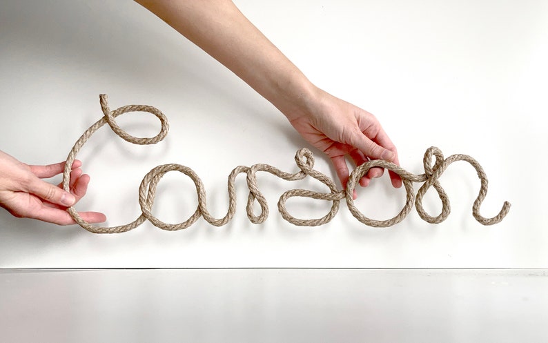 May include: A decorative wall sign formed from thick, natural-colored twisted jute rope, spelling out the name Carson in a continuous, flowing cursive script. The rope is textured and tan, creating a structured, legible word display.