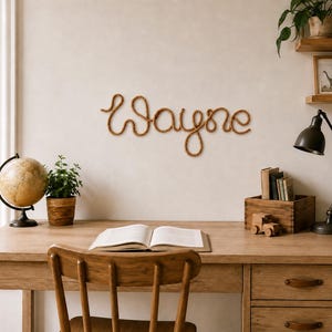 May include: A wall-mounted sign featuring the name Wayne written in a continuous, cursive script made from a thick, brown, twisted rope material. The letters are connected in a single flowing line, creating a decorative piece of wall art.