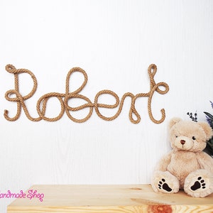 May include: A wall-mounted name sign spelling the name Robert, crafted from thick, twisted brown rope. The letters are connected in a continuous cursive script. A plush beige teddy bear and a small potted plant are positioned below the sign.