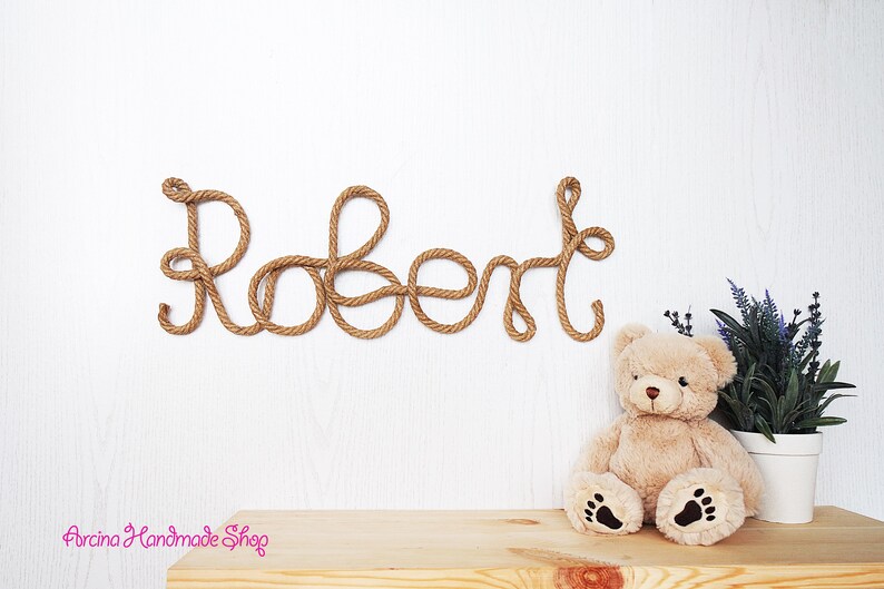 May include: A wall-mounted name sign spelling the name Robert, crafted from thick, twisted brown rope. The letters are connected in a continuous cursive script. A plush beige teddy bear and a small potted plant are positioned below the sign.